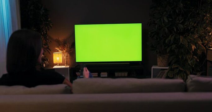 A young woman sitting on couch in the cozy living room watching a television whileresting relaxing after hard long day at work at home green screen mockup.