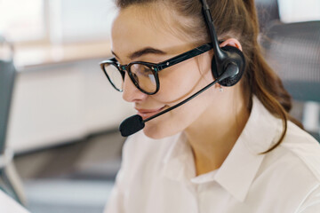 A close-up of a young woman wearing glasses and a headset, engaged in a conversation in a modern office.
