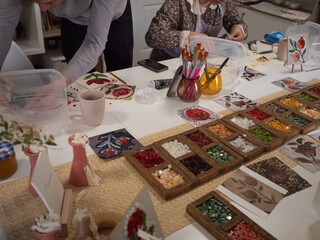 Mosaic Workshop: Horizontal shot of two pairs of hands - male and female - working at the table with boxes of colourful vitrage glass, pliers, samples of ancient floral patterns, and other tools.