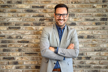 Portrait of young businessman with arms crossed posing confidently against brick wall in modern office