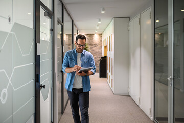 Smiling male financial advisor in eyeglasses analyzing reports while walking in office corridor