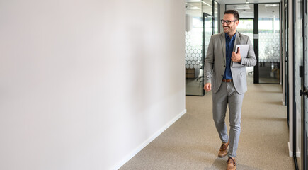 Full length of happy male leader with laptop looking away and walking by white wall in office...