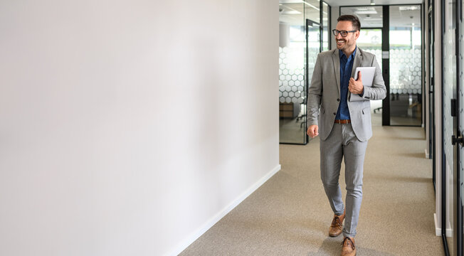 Full length of happy male leader with laptop looking away and walking by white wall in office corridor
