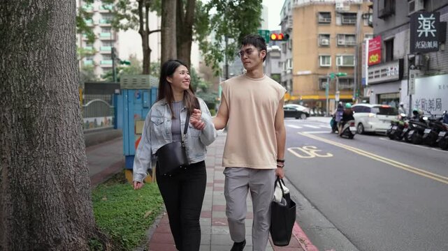 A young Taiwanese couple in their twenties walking through the streets of Xinyi District, Taipei City at noon in spring. The man is wearing a sleeveless top, and the woman is wearing a denim jacket.