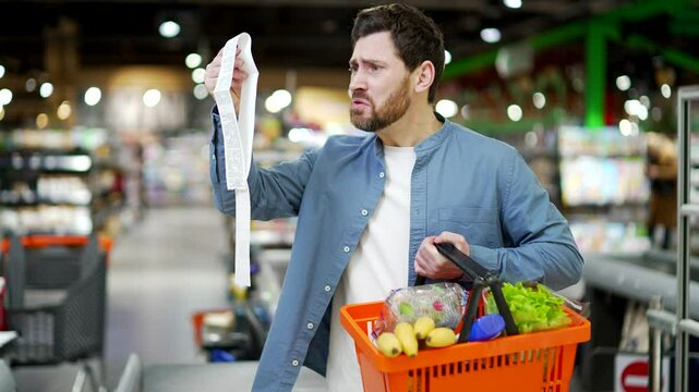 Unhappy customer holding a long grocery receipt check. Man feeling shocked about rising grocery high prices inflation. Gets shocked looking at check after shopping Male Checking Shopping Bill