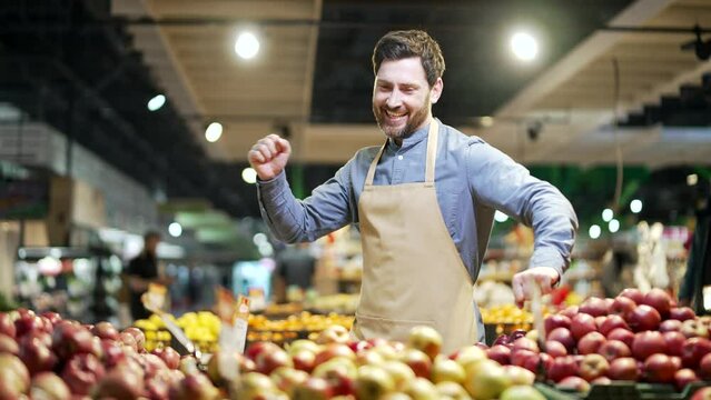 Happy positive greengrocer dancing near counter with fruits and vegetables. A male salesman in grocery store, market or supermarket is having fun. Cheerful worker in apron or a small business owner
