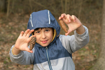 Child with a blue hooded jacket holding pine cones, smiling with excitement. Concept: Reflecting the significance of teaching children about nature and fostering a sense of wonder and respect for the