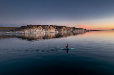 Young woman rides on stand up paddle sup board at calm sunset lake against white cliff and blue sky