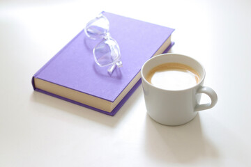 A purple mock up book with transparent glasses and a white cup of coffee on white table. Free copy space for text