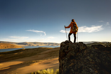 Woman hiker with hiking poles and backpack is standing on big rock against sunset desert, blue sky and lake