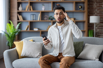 Obraz premium Portrait of a young worried Indian man at home, sitting on the sofa, holding the phone, holding his head with his hand, looking at the camera in shock