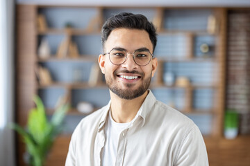 Fototapeta premium Close-up photo of a young Muslim man in glasses and a shirt sitting at home and smiling at the camera
