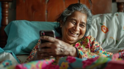 a senior smiling indian woman lying in bed, looking at her cell phone