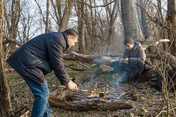 Two men make a campfire for cooking in the forest, father and son sitting on a log while hiking and outdoor activities, early spring landscape