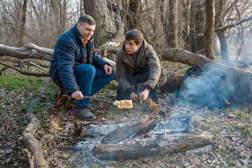 Two men make a campfire for cooking in the forest, father and son sitting on a log while hiking and outdoor activities, early spring landscape