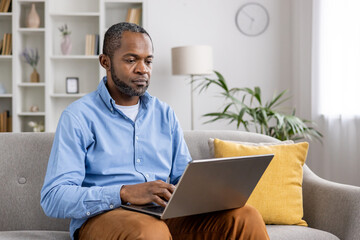 A man working from home on his laptop while sitting on a comfortable sofa in a well-decorated living room. The setting is relaxed, promoting productivity and focus.