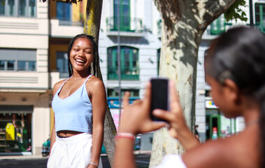 Young black woman taking pictures of model smiling with mobile phone while posing on the street. Photography and travel concept