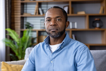 A serious man in a blue shirt is sitting on a sofa in a modern living room. The background features...