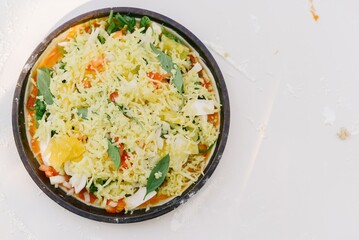 A close-up shot of a pizza with tomato sauce, mozzarella, basil, onions, orange, and cheese ready to be baked. The pizza is on a brown rim with a white background.