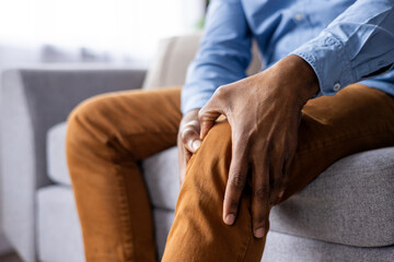 Close-up shot of a person holding their knee, indicating pain or discomfort. The individual is seated on a sofa,. Ideal for topics related to health, knee pain, and discomfort. © Liubomir