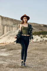 stylish woman in plaid shirt and hat strolls confidently through the desert landscape with hands in pockets