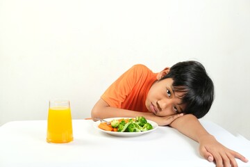 A boy with a tired expression rests his head on the table next to a plate of vegetables and a glass...