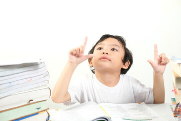 A young boy, wearing a white shirt, sits between stacks of books, looking up with both index fingers pointing upwards. His expression is thoughtful and curious.