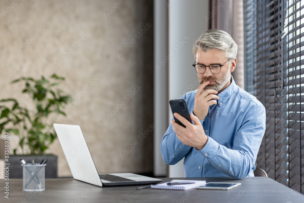 Wall mural Mature businessman with glasses looks at his smartphone and laptop, working in a modern office. The setting includes a notebook, pen, and a plant in the background.