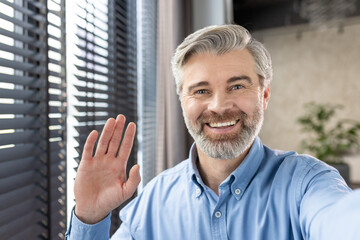 Happy man with gray hair and beard waving at the camera while having a video call in a well-lit modern office. He is wearing a blue shirt and smiling warmly.