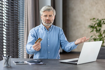 Senior man looking frustrated while holding a credit card in one hand and gesturing with the other hand at home. He is sitting in front of a laptop, facing an online payment issue.