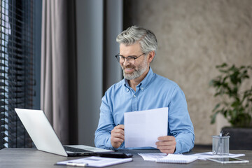 A smiling mature businessman is seen working at his desk in a modern office. He is holding documents and looking at a laptop, indicating productivity and satisfaction in his work environment.