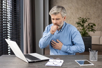 Middle-aged man working from home is seen coughing and holding his chest while seated at a desk...
