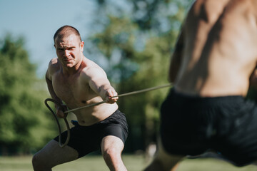 Two fit, shirtless men participating in an intense tug of war exercise in a sunny park setting, showcasing strength and determination.