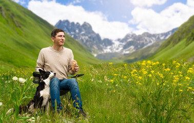 A man sits on a camp chair during a rest stop and eats something while his Bored Collie sits next to him. Active recreation with a dog in nature in the mountains. Travel with dog