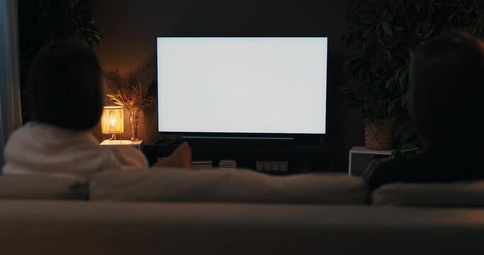 Two friends are talking last gossips weekends concept. Happy women sitting in the living room watching tv news movie white screen mockup television in cozy living room over the shoulder shot.