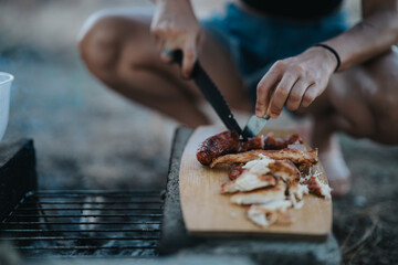 Close-up of hands cutting and preparing grilled meat and sausages on a wooden board during an outdoor barbeque.