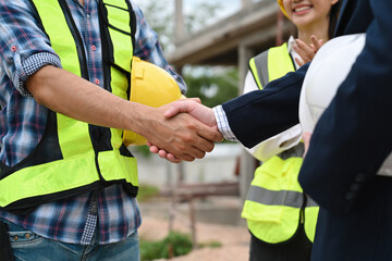 Close-up of a contractor and a businessman shaking hands at a construction site, with a female worker clapping in the background, representing a successful agreement