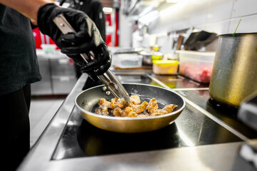 A chef’s hands in black gloves cooking shrimp in a stainless steel pan over a stove.