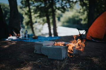 Preparing a barbeque fire at a mountain picnic by the river, with an orange tent and a scenic view under the shade of trees.