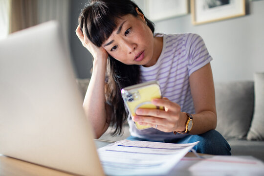 Concerned Asian woman holding smartphone looking at laptop on sofa