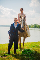 A man and a woman in historical costumes on the shore of a lake. The man leads a horse on which the woman sits.