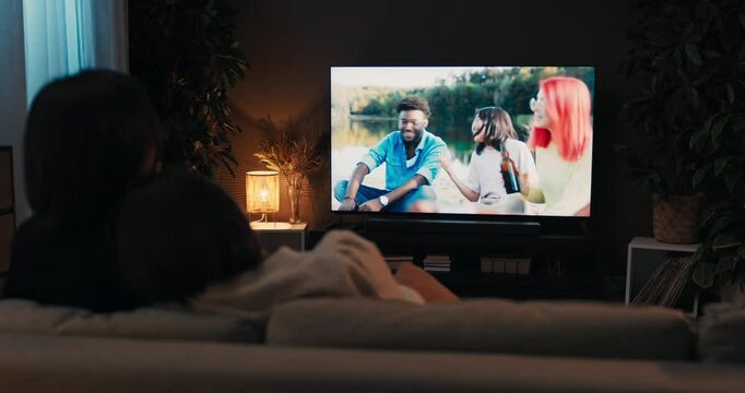 Brunette young women couple lesbian sitting in the living room watching tv resting at the weekends afterlong had week. Realtionship concept. Over the shoulder shot.