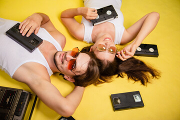 A woman and a man in sunglasses are lying on a yellow background. An old tape recorder and video...