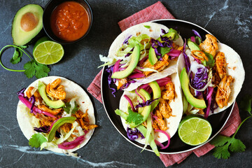 Chicken street tacos with avocado, cabbage slaw and pickled red onions. Top down view table scene on a dark stone background.