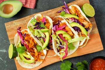 Chicken street tacos with avocado, cabbage slaw and pickled red onions. Above view table scene with platter on a dark stone background.