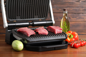Electric grill with raw meat, oil and vegetables on wooden table