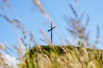 A cross stands tall on a hilltop, with blue sky in the background and blurred field grass in the...