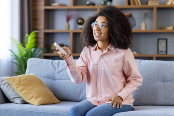 A woman with glasses and curly hair sits on a couch holding an air conditioning remote. She looks...