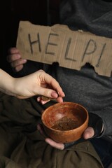 Woman giving coins to poor homeless man with bowl of donations and help sign on dark background, closeup