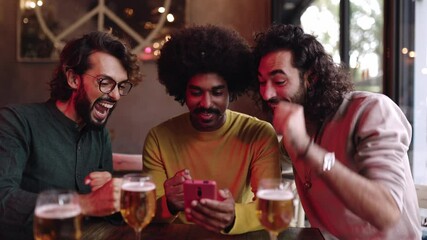 Three young male friends betting online celebrating success while using mobile phone sitting on table at bar, drinking beer and having fun. Slow motion 4K video.